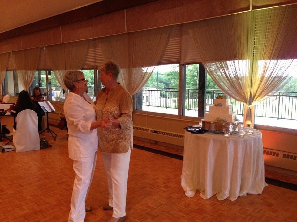 Two white women slow dance together indoors. Next to them is a table with a large wedding cake.