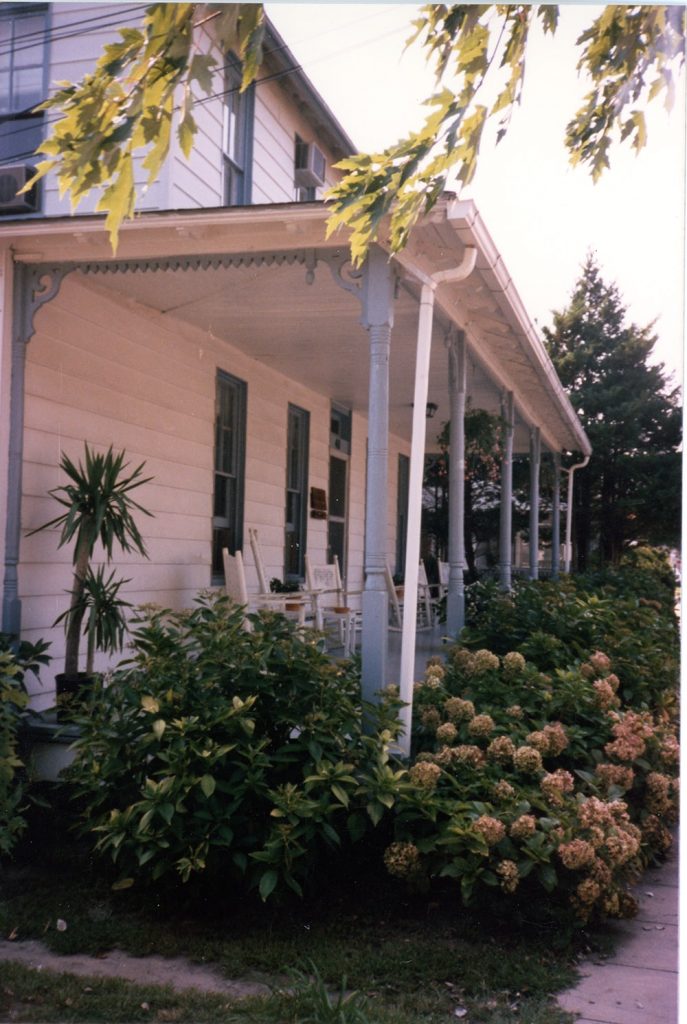 A porch with decorative moldings is seen from the side. Lush greenery is seen in front of the porch.