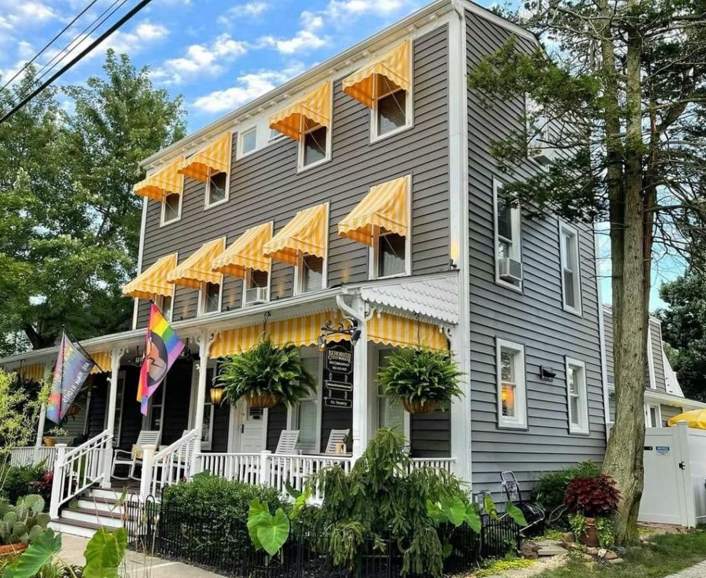 A three-story, gray house with yellow-striped awnings and a white porch is seen with two pride flags.