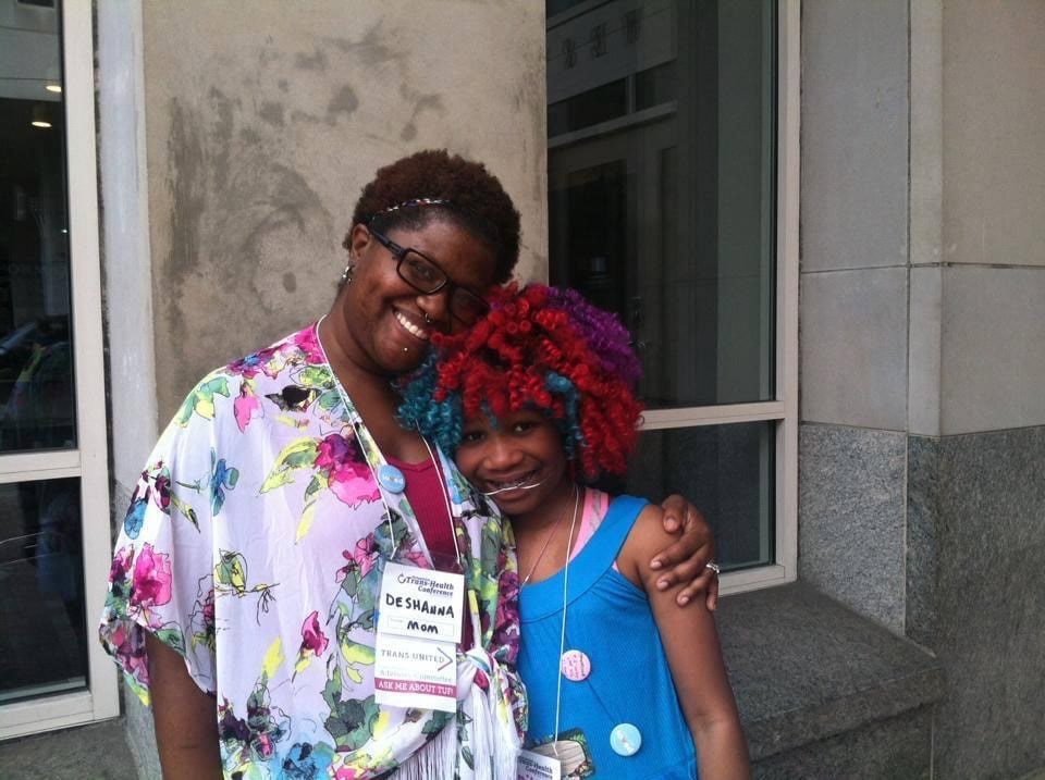 A Black woman with short hair smiles and puts her arm around her daughter who is wearing a purple, hot pink, and teal wig.