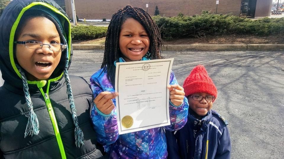 Three Black children look at the camera with the girl in the center holding a birth certificate.