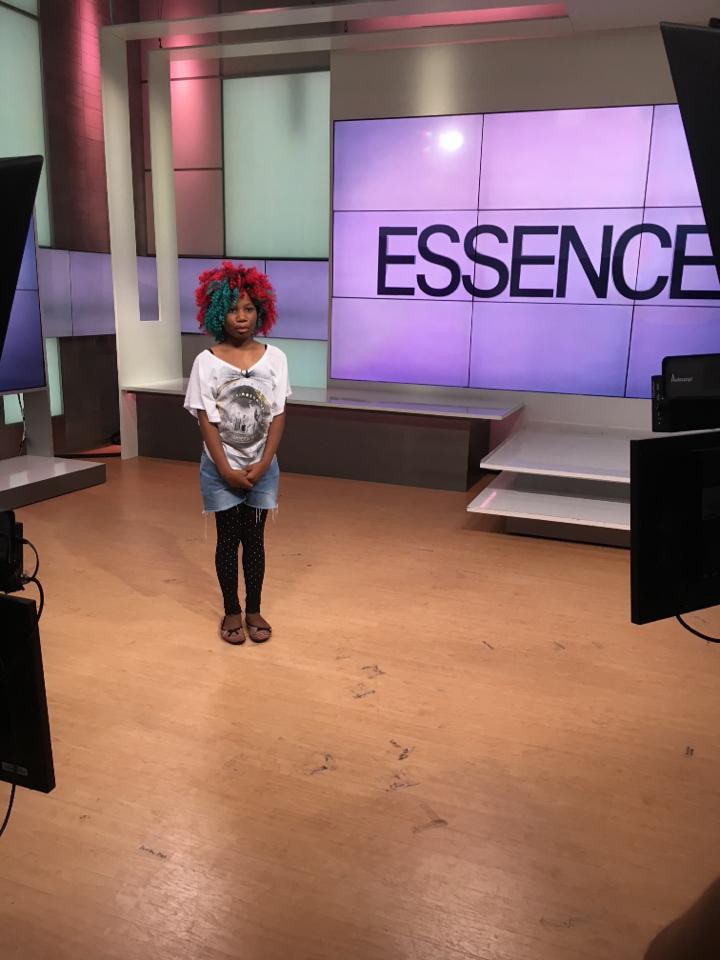 A Black girl wearing a purple, hot pink and teal wig is seen being interviewed by Essence magazine.
