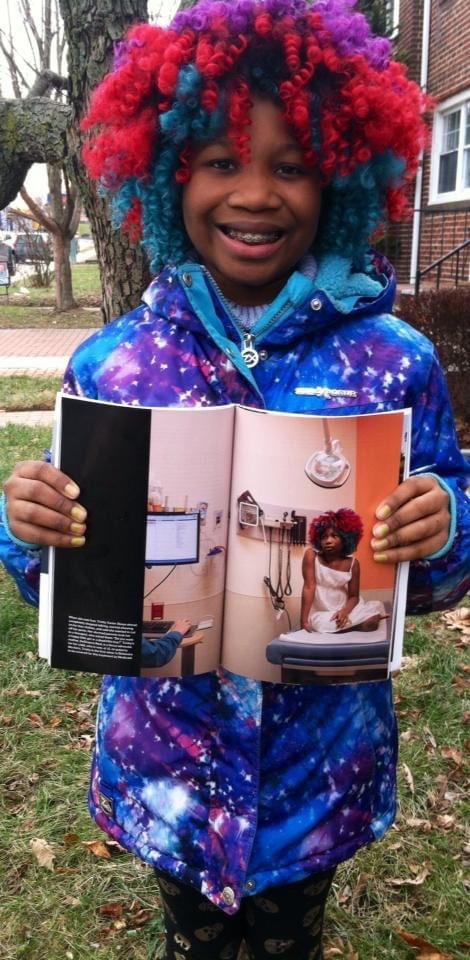 A Black girl standing outside in a purple, hot pink and teal wig shows the viewer a two-page photograph of herself sitting on a hospital bed.