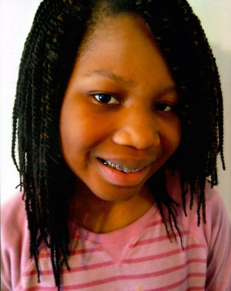 A smiling Black girl with medium-length, black hair. She has braces and is wearing a striped pink shirt.