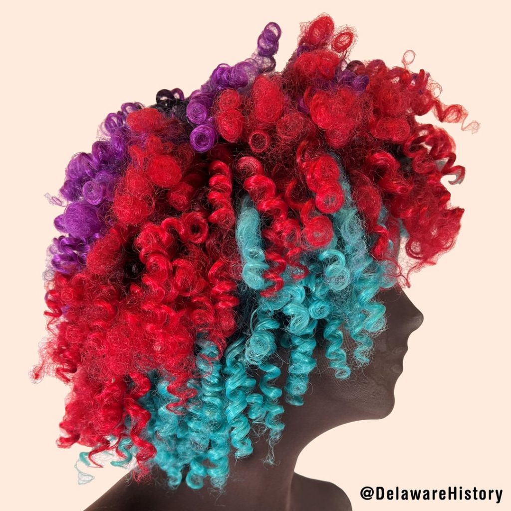 A purple, hot pink and teal wig is displayed on a dark-skinned mannequin.