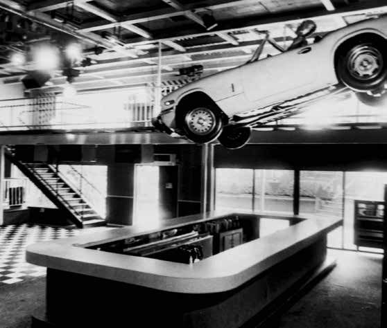 A black and white photograph of a convertible with a mannequin at the wheel floating above a bar.