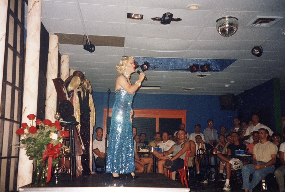 A white drag queen performs on stage in front of patrons, wearing a sequined blue dress with a blonde wig.