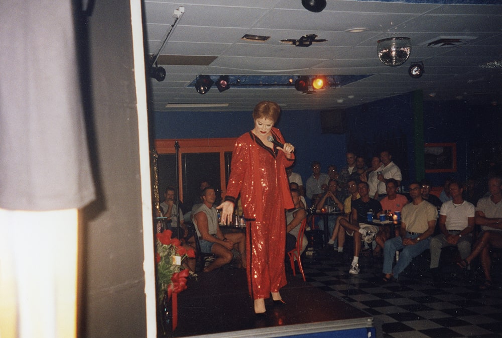 A white drag queen performs on stage in front of patrons, wearing a sequined red pants suit with a red wig.