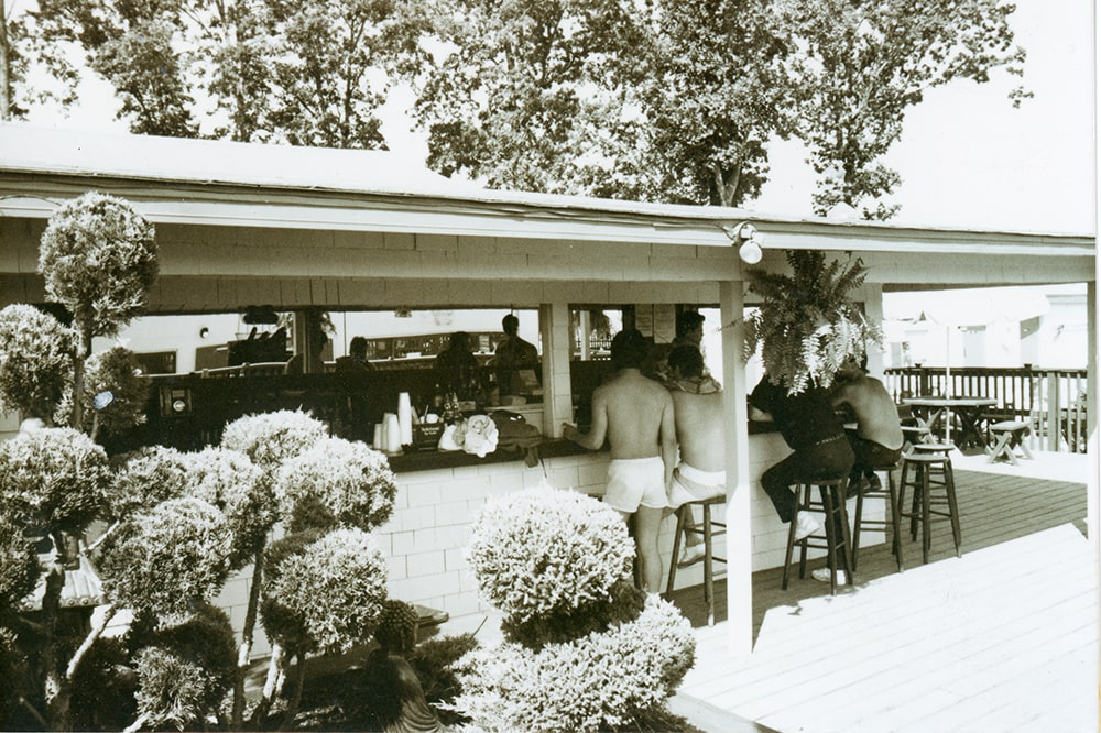 3 shirtless men and 1 fully dressed man are seen from behind sitting and standing at the pool bar at the Renegade.