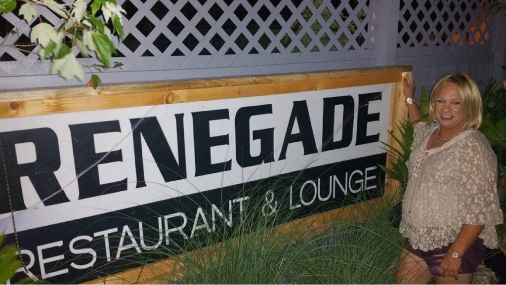 A white woman with blonde hair poses with the black and white sign reading “Renegade Restaurant & Lounge.”