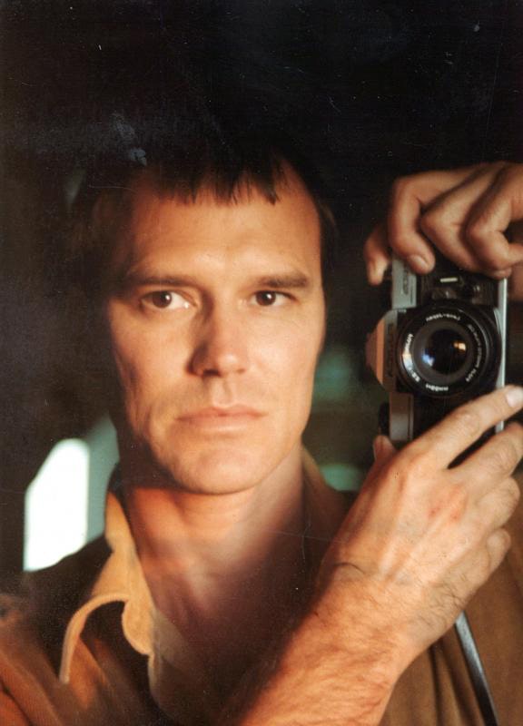 A white man with short light-brown hair takes a self-portrait in the mirror with a film camera.