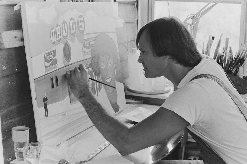 A black and white photograph of a white man wearing a white t-shirt and overalls working on a painting of a Black woman in front of a drug store.