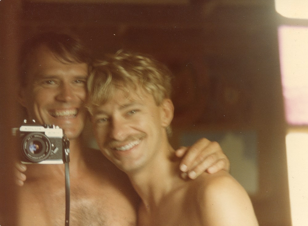 A shirtless white man with short brown hair takes a photograph in the mirror with another shirtless white man with blonde hair.