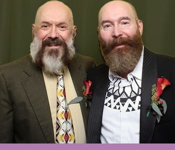 Two light-skinned men with full beards and mustaches smile for the camera. Both men are wearing suit jackets with red flowers pinned to the lapels.