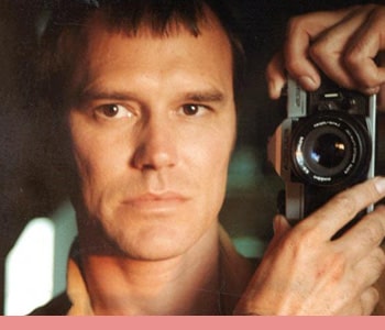 A white man with short light-brown hair takes a self-portrait in the mirror with a film camera.