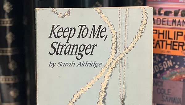 A paperback copy of the novel “Keep To Me, Stranger,” is displayed. The cover depicts two necklaces, a beaded necklace and the star of David