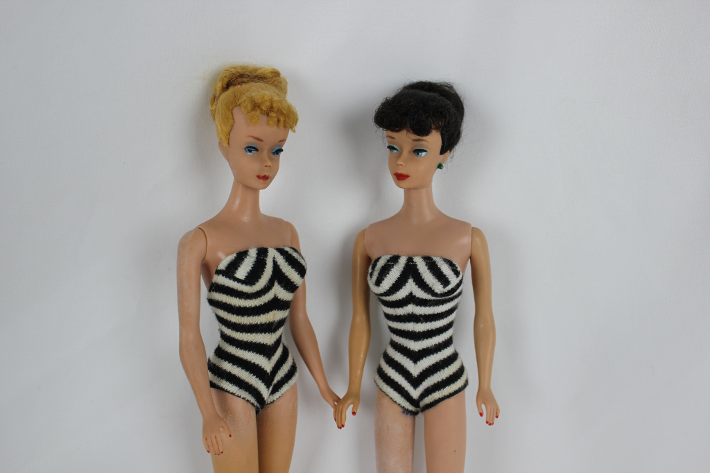 Two original Barbie dolls, both Caucasian, one with blonde hair and the other with black hair, wearing black and white striped swimsuits.