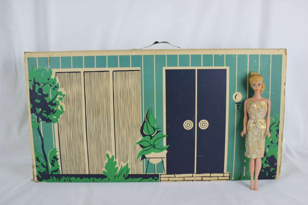 : Cardboard exterior of Barbie's dream home doll house, showing the green printed paneling, door, and windows, with Barbie standing in front of it wearing a gold cocktail dress