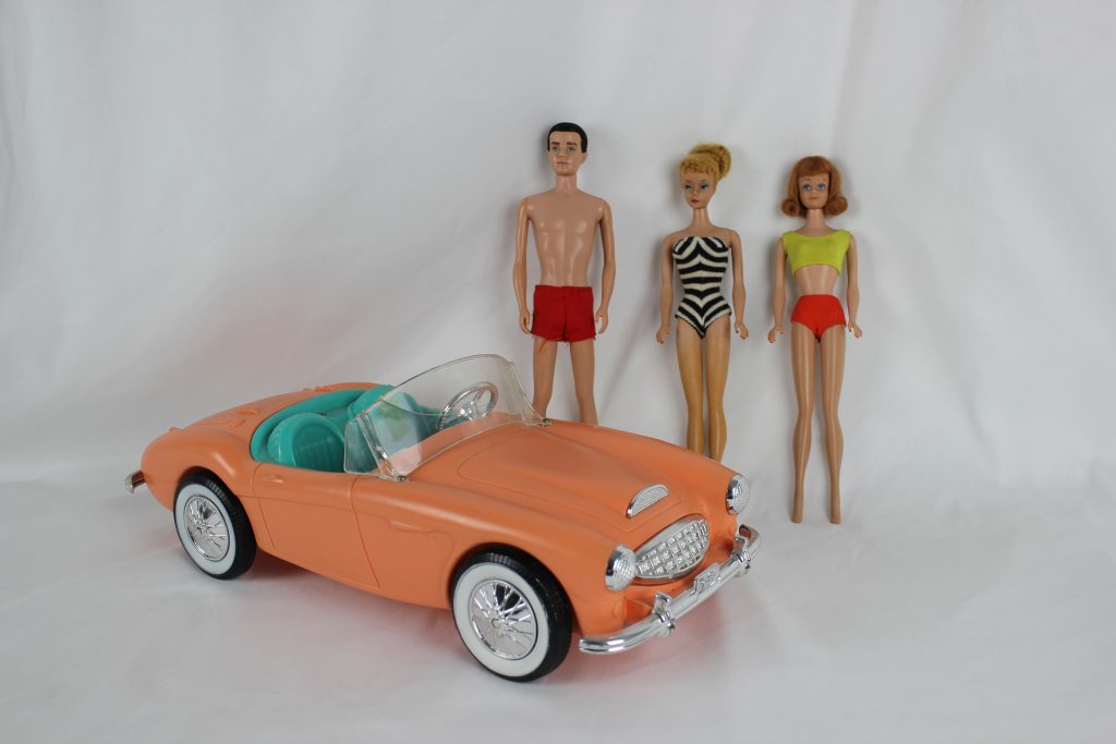 Barbie, Ken, and Midge standing beside Barbie's pink toy convertible