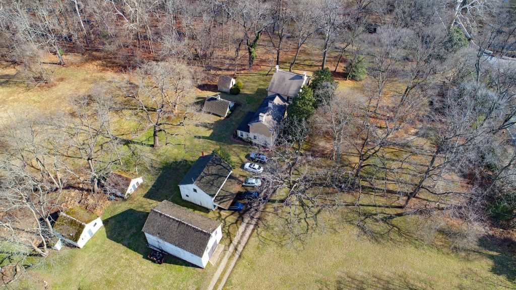 Aerial view of historic home, outbuildings and cars in a wooded clearing.