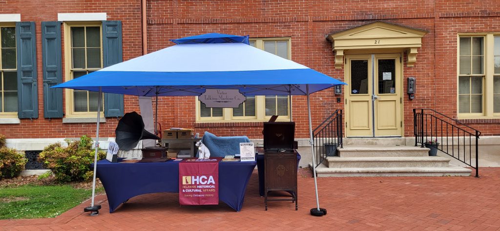 A blue and white covered display table with an HCA logo features victrolas and a statue of the white dog with black ears, Nipper, in front of the Old State House.