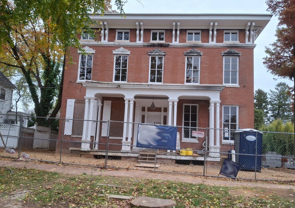 Exterior front view of three-story brick home with four white columns behind a chain-link fence and port-o-san at right under renovation.