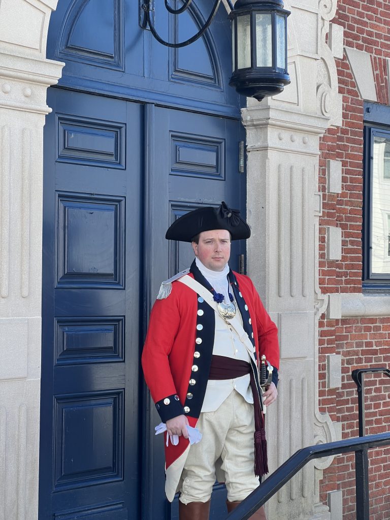 White male wearing traditional British Redcoat attire with black hat and sword on his hip stands in front of brick building with blue door and black iron lighting fixture.