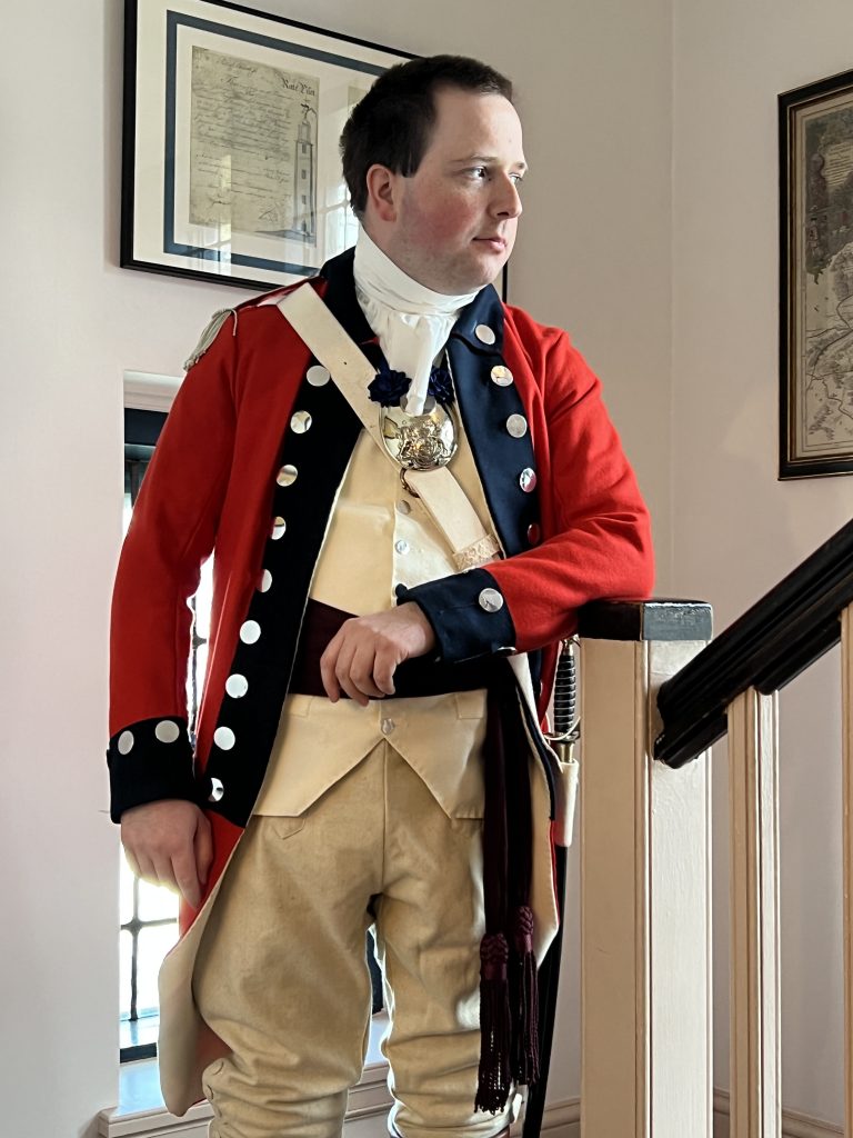 While male in traditional British Redcoat attire with white shirt, beige vest, pants and sash. buttons along the blue rimmed coat edges and cuffs, sword on his hip, posing on stairs.