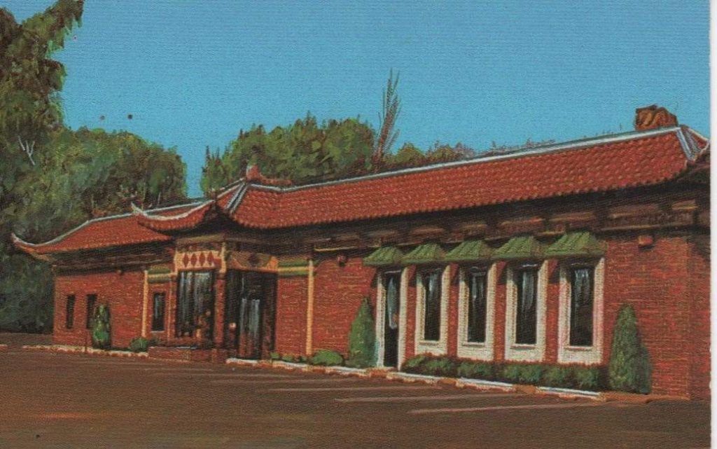 Building with a red tile roof and curved eaves, upturned roof corners that were adorned with dragons at the ridge line. There is a centrally placed glass vestibule with side door entries and elongated fixed windows highlighted by wide surrounds topped with green overhanging hooded eaves. 