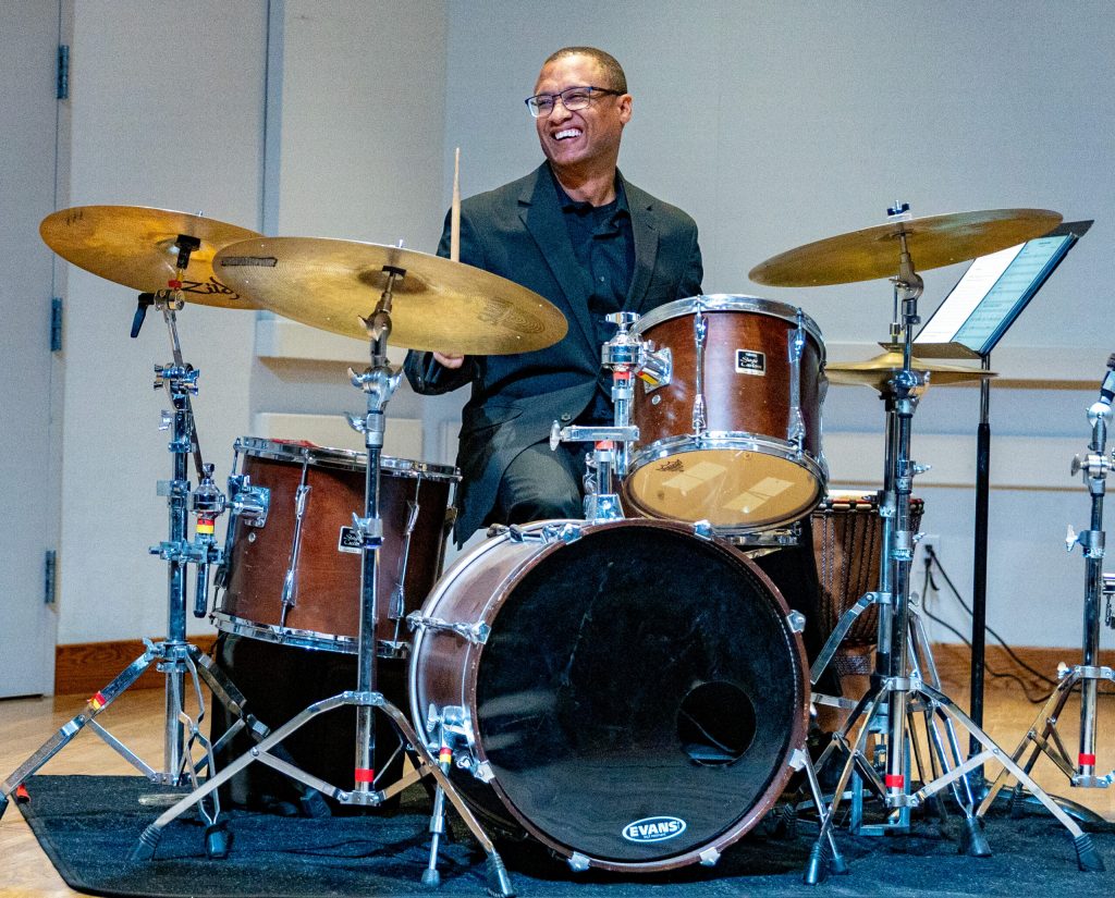 African American composure plays drums while smiling broadly.