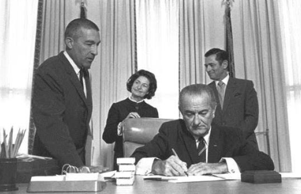 Black and white image of President Johnson, seated at his desk, flanked by two men and woman, as he signs a document.